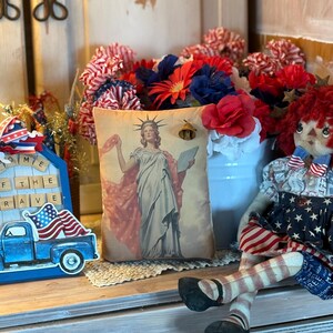 May include: A patriotic display featuring a cushion with a Statue of Liberty image, a blue truck with an American flag, and a rag doll. Red, white, and blue floral arrangements and decorative accents complete the festive arrangement.