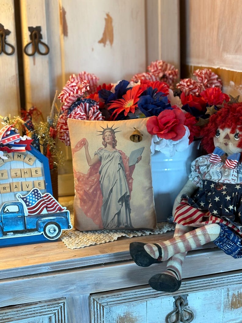 May include: A patriotic display featuring a cushion with a Statue of Liberty image, a blue truck with an American flag, and a rag doll. Red, white, and blue floral arrangements and decorative accents complete the festive arrangement.
