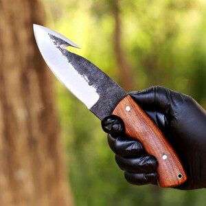 May include: A handcrafted knife with a wooden handle and a curved blade. The blade has a hook at the tip and a dark, textured finish. The handle is a rich brown color, secured with silver rivets.