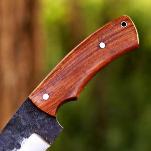 May include: Close-up of a handcrafted knife. The handle is made of dark, polished wood, secured with three silver rivets. The blade is dark, with a textured, forged appearance. The knife is designed for cutting and chopping.