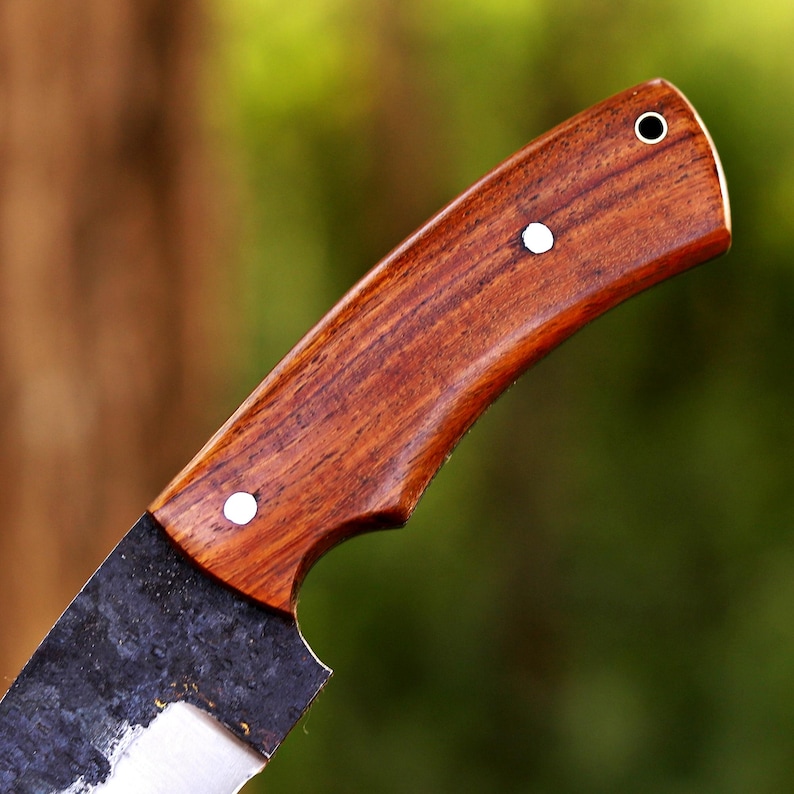 May include: Close-up of a handcrafted knife. The handle is made of dark, polished wood, secured with three silver rivets. The blade is dark, with a textured, forged appearance. The knife is designed for cutting and chopping.