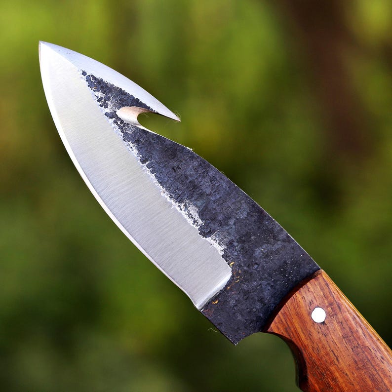 May include: Close-up of a handcrafted knife with a polished steel blade and a dark, textured edge. The handle is made of wood and features a single rivet. The knife has a unique hook-shaped tip.