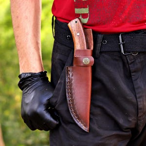 May include: A wooden-handled knife in a brown leather sheath, secured to a black belt. The sheath has decorative stitching along the edge and a circular emblem. The knife handle is light brown, and the sheath is a darker shade of brown.