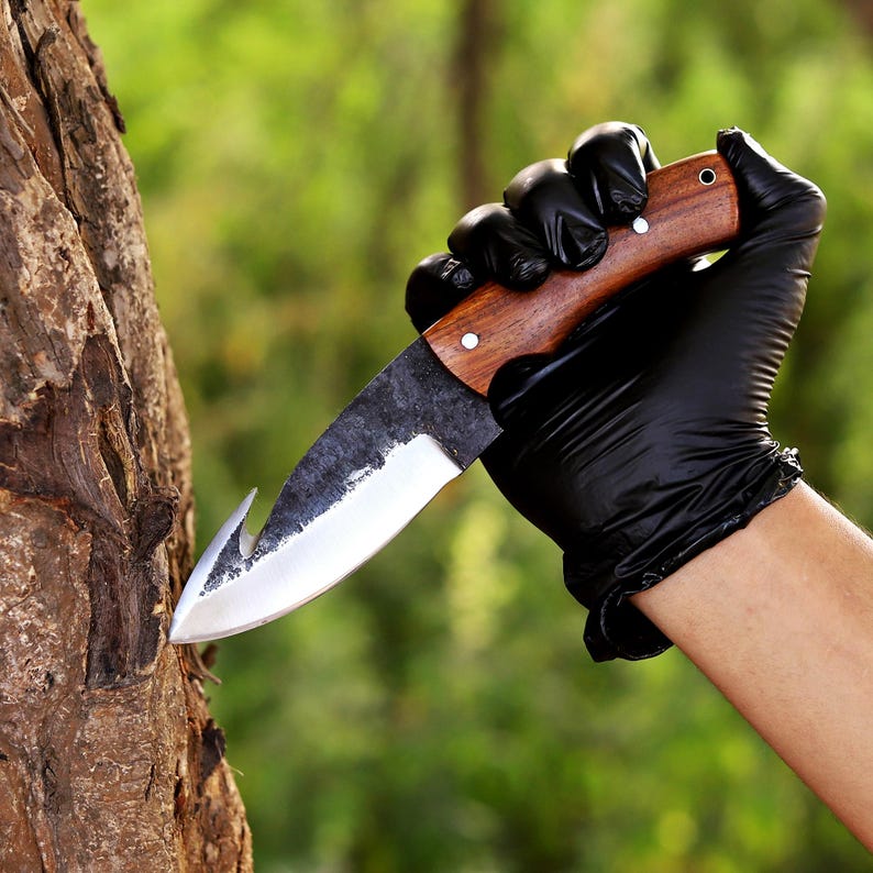 May include: A handcrafted knife with a wooden handle and a curved blade. The blade has a hook-like tip and a dark, textured finish. The handle is made of wood and has a smooth, polished finish. The knife is held by a person wearing black gloves.