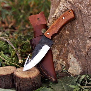 May include: A handcrafted knife with a wooden handle and a curved blade, resting in a brown leather sheath. The knife has a rustic, forged appearance, with a sharp edge and a hook at the tip. The sheath has a belt loop and decorative metal accents.