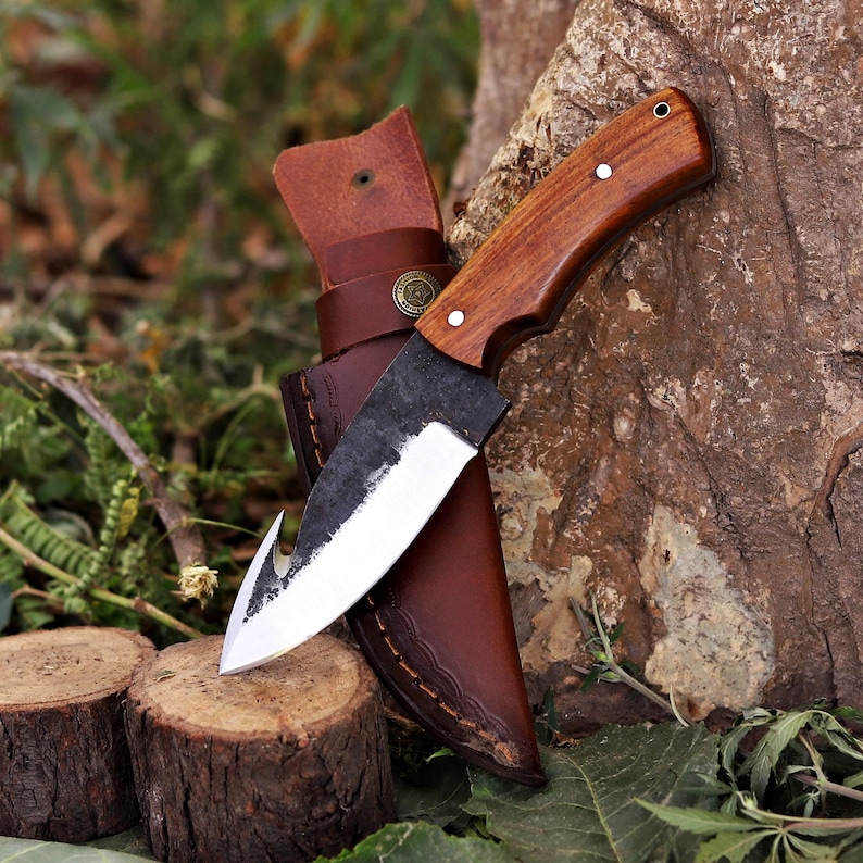 May include: A handcrafted knife with a wooden handle and a curved blade, resting in a brown leather sheath. The knife has a rustic, forged appearance, with a sharp edge and a hook at the tip. The sheath has a belt loop and decorative metal accents.