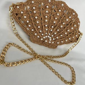 Puede incluir: Bolso de mano en forma de concha, tejido en material marrón, adornado con perlas blancas. Cuenta con una correa de cadena dorada. El diseño evoca un ambiente playero o costero, ideal para llevar lo esencial.