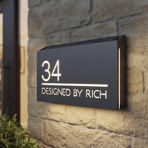 Puede incluir: Un letrero rectangular de número de casa en negro mate que muestra el número 34 y el texto DESIGNED BY RICH en letras blancas iluminadas. El letrero tiene una barra de luz horizontal y una tira de luz vertical en el lado derecho, montado en una pared de piedra.