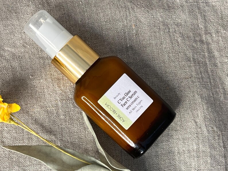 Peut inclure: Une bouteille en verre marron de s&eacute;rum pour le visage avec une pompe blanche et dor&eacute;e. L'&eacute;tiquette indique "C You Glow Face C Serum with Vitamin C" et "ACRE+BLOOM". La bouteille est sur une surface de couleur neutre.
