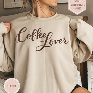 May include: A sand-colored sweatshirt with the words "Coffee Lover" in a brown script. The long-sleeved top is made of a soft material. A small, round, sand-colored sticker with the word "SAND" is visible.