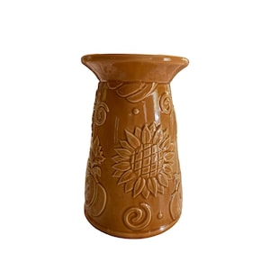 May include: A brown ceramic candle holder with a sunflower and other embossed designs. The candle holder has a wide top and a flared base. The sunflower is the central design element, with other embossed elements including leaves and swirls.