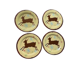 May include: Four round plates with a light yellow center and a brown rim. Each plate features a brown reindeer silhouette and blue and green snowflake designs. The plates are arranged on a white background.