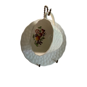 May include: A white teacup and saucer set with a textured surface. The inside of the cup features a floral design with fruits and flowers. The set is suspended by a brown rope.