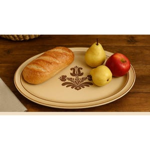 May include: An oval beige serving platter with a brown floral design, a loaf of bread, two yellow pears, and a red apple. The platter has a brown rim and is set on a wooden surface.