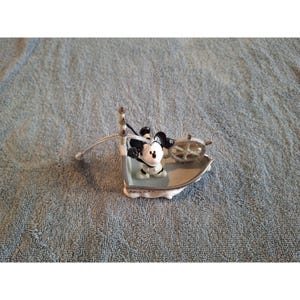 Pode incluir: Uma pequena figura decorativa do Mickey Mouse num barco. O barco &eacute; prateado com uma borda branca e um pequeno volante. Mickey est&aacute; usando sua roupa cl&aacute;ssica e parece estar dirigindo o barco. O enfeite est&aacute; em uma toalha azul.