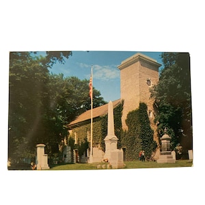 May include: Vintage postcard featuring a stone church with a tall tower and an American flag. The church is covered in ivy and surrounded by trees. A white obelisk and other monuments are in the foreground. The sky is blue with some clouds.