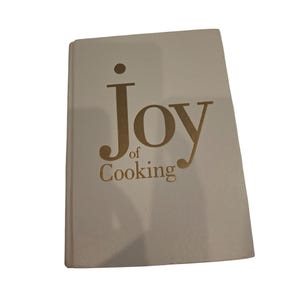 May include: A hardcover book titled Joy of Cooking with a white cover and gold lettering. The title is printed in a large, stylized font on the front cover. The book is rectangular with a clean, smooth surface and a visible spine on the left side.