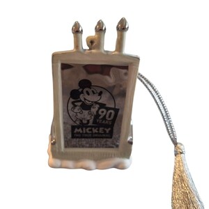 Pode incluir: Um ornamento retangular com Mickey Mouse celebrando 90 anos. O ornamento tem um esquema de cores prateado e branco com tr&ecirc;s estruturas prateadas semelhantes a chamin&eacute;s no topo. Um cord&atilde;o e borla prateados s&atilde;o anexados para pendurar.