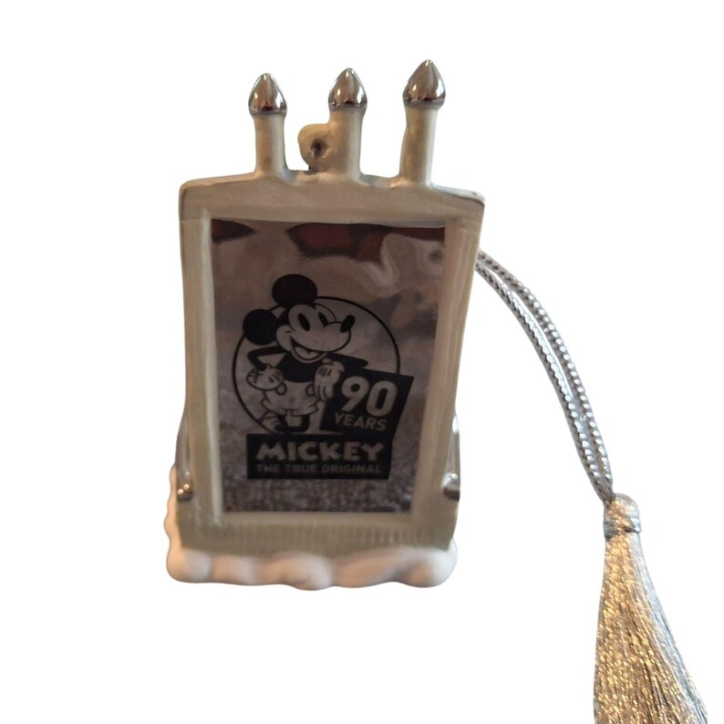Pode incluir: Um ornamento retangular com Mickey Mouse celebrando 90 anos. O ornamento tem um esquema de cores prateado e branco com tr&ecirc;s estruturas prateadas semelhantes a chamin&eacute;s no topo. Um cord&atilde;o e borla prateados s&atilde;o anexados para pendurar.