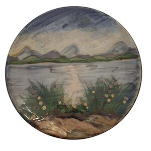 May include: A round ceramic plate featuring a hand-painted landscape scene. The design depicts a calm lake reflecting light, surrounded by green grassy banks with small yellow flowers in the foreground and a range of mountains under a cloudy sky.