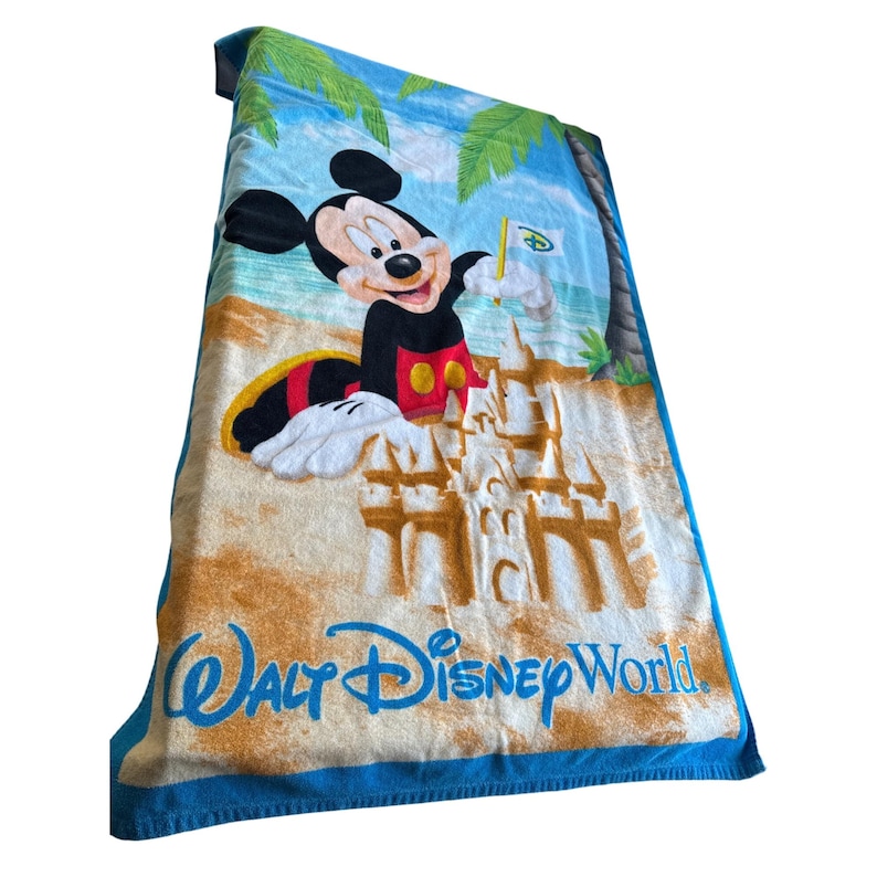 May include: A blue beach towel featuring Mickey Mouse on a beach with a sandcastle and palm trees. The towel has the words "Walt Disney World" in blue script. Mickey Mouse is wearing his signature red shorts and holding a flag.