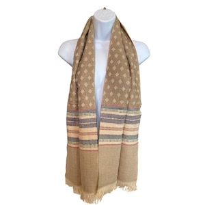 May include: A tan scarf with a geometric diamond pattern and striped accents in red, green, and blue. The scarf has a fringed edge and is displayed on a white mannequin.