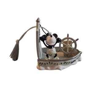 Pode incluir: Estatueta do Mickey Mouse num barco azul claro e branco, com um volante prateado. Mickey usa sua roupa cl&aacute;ssica. O barco &eacute; decorado com detalhes prateados e um cord&atilde;o com borla.