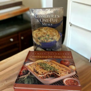 May include: Two hardcover cookbooks stacked on a surface. The top book is titled 125 Best Casseroles & One-Pot Meals by Rose Murray, featuring a blue dish. The bottom book is titled All Time Family Favorites Casseroles & One-Dish Meals.