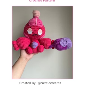 May include: A crocheted amigurumi toy in the shape of a sad girl holding a purple hammer. The toy is pink with white eyes and a pink and white hat. The toy is being held in a hand.