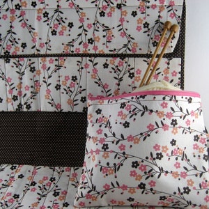 May include: A fabric organizer featuring a white background with a floral pattern of pink, peach, and dark brown flowers on thin black stems. The organizer includes a matching zippered pouch with a pink zipper and two wooden knitting needles.