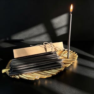 May include: A collection of thin black taper candles resting on a gold-colored feather-shaped tray. One candle is lit and stands upright, while the others are bundled together with twine and a rectangular brown paper sleeve.