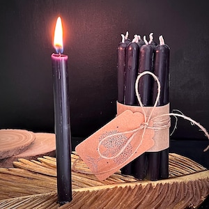 Peut inclure: Une bougie noire allumée avec une flamme orange vif est à côté d'un paquet de bougies noires. Les bougies sont attachées avec de la ficelle et une étiquette marron avec un motif d'arbre. Les bougies sont sur un plateau doré en forme de feuille.