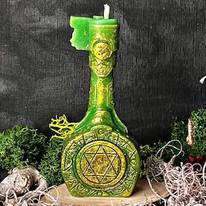 Könnte beinhalten: Eine grüne Kerze in Flaschenform mit einem goldenen Stern-Design. Die Kerze hat detaillierte Muster und steht auf einem Holzsockel mit grünem Moos. Das Design der Kerze ist aufwendig und dekorativ.