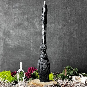 May include: Black candle shaped like a broom with a wooden handle. The candle is sitting on a wooden base surrounded by moss and greenery.