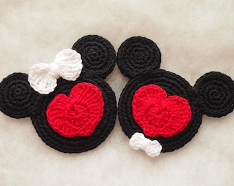 May include: Two crocheted black Mickey Mouse-shaped coasters, each with a red heart and a white bow. One bow is on the top, and the other is on the bottom. The coasters are handmade.