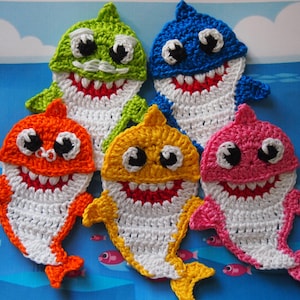 May include: Five crocheted shark toys in different colors: green, blue, orange, yellow, and pink. Each shark has white crocheted details, black eyes, and a red mouth.