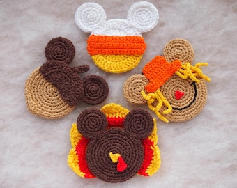May include: Four crocheted coasters featuring fall-themed designs. The coasters are shaped like Mickey Mouse heads and feature a candy corn, a scarecrow, a turkey, and a brown acorn.