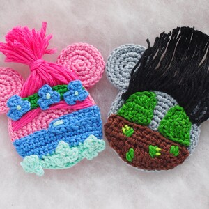 May include: Two crocheted character ornaments, one pink with blue and green accents and the other gray with green and brown accents.