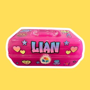 May include: A pink, glittery, rectangular carrying case with a handle. The name "LIAN" is displayed in blue and white block letters. The case is decorated with stickers of hearts, stars, and a diamond.