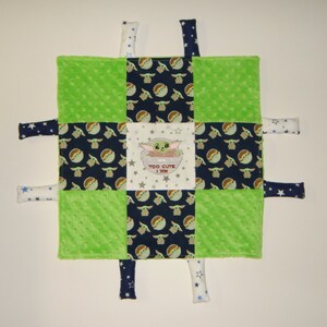 K&ouml;nnte beinhalten: Eine gr&uuml;ne, blaue und wei&szlig;e Babydecke mit einem quadratischen Muster. Das mittlere Quadrat zeigt ein gesticktes Bild eines gr&uuml;nen Charakters mit dem Text "TOO CUTE I AM". Die Decke hat sechs Stoffb&auml;nder, die an den Ecken und Seiten befestigt sind.