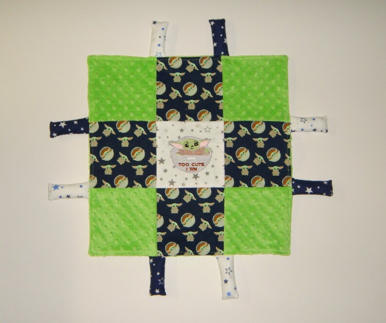 K&ouml;nnte beinhalten: Eine gr&uuml;ne, blaue und wei&szlig;e Babydecke mit einem quadratischen Muster. Das mittlere Quadrat zeigt ein gesticktes Bild eines gr&uuml;nen Charakters mit dem Text "TOO CUTE I AM". Die Decke hat sechs Stoffb&auml;nder, die an den Ecken und Seiten befestigt sind.