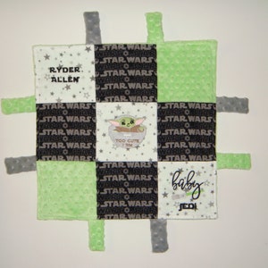 K&ouml;nnte beinhalten: Eine gr&uuml;ne, schwarze und wei&szlig;e Babydecke mit Star Wars-Motiv. Die Decke hat ein quadratisches Design mit einer Figur aus dem Star Wars-Franchise in der Mitte. Die Decke hat an jeder Ecke Etiketten. Die Decke tr&auml;gt den Text "Ryder Allen" und "Baby Jedi".