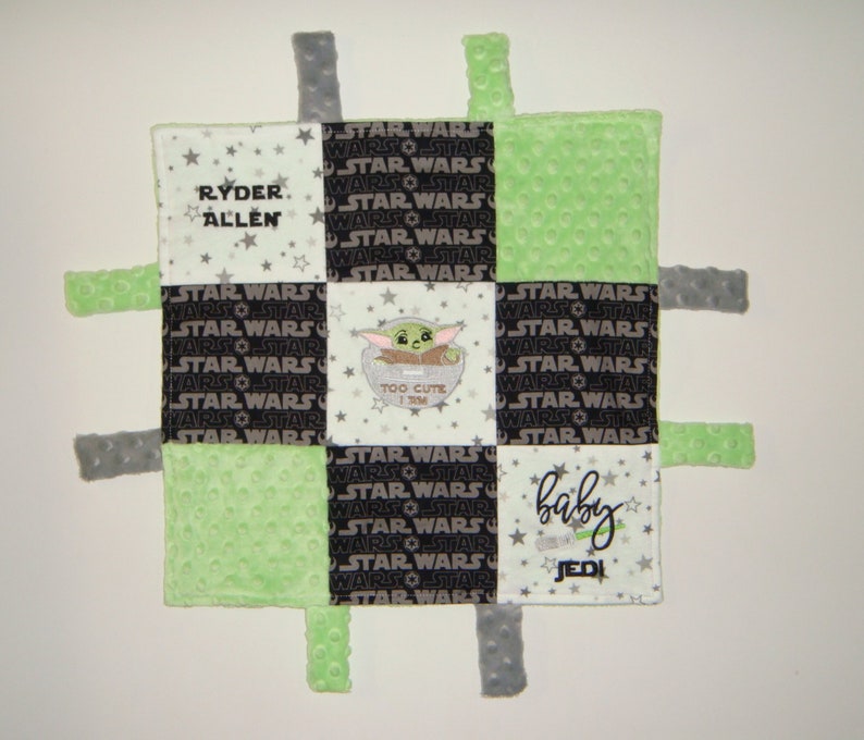 K&ouml;nnte beinhalten: Eine gr&uuml;ne, schwarze und wei&szlig;e Babydecke mit Star Wars-Motiv. Die Decke hat ein quadratisches Design mit einer Figur aus dem Star Wars-Franchise in der Mitte. Die Decke hat an jeder Ecke Etiketten. Die Decke tr&auml;gt den Text "Ryder Allen" und "Baby Jedi".