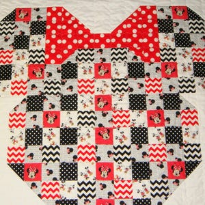 May include: A patchwork quilt in the shape of a Minnie Mouse bow and top. The quilt features a red polka dot bow and a patchwork design of red, black, white, and gray squares with Minnie Mouse images and zig-zag patterns.