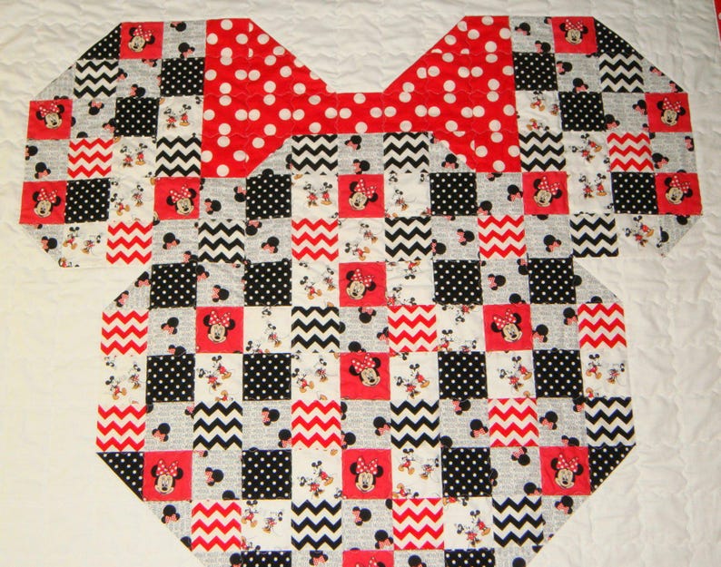 May include: A patchwork quilt in the shape of a Minnie Mouse bow and top. The quilt features a red polka dot bow and a patchwork design of red, black, white, and gray squares with Minnie Mouse images and zig-zag patterns.