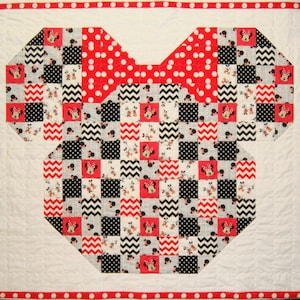 May include: A handmade quilt featuring a Minnie Mouse design. The quilt is shaped like Minnie's silhouette, with a large red polka dot bow. The body is a patchwork of black, white, and red squares with Minnie Mouse images. The border is white with a red polka dot trim.