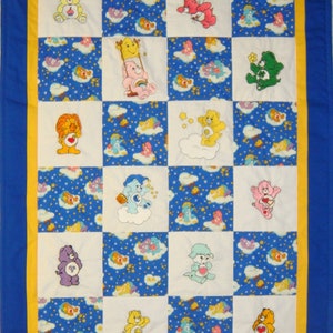 May include: A blue and white patchwork quilt with a yellow border. The quilt is made up of squares with different Care Bear characters on each square. The characters are all smiling and have different colored fur and accessories.