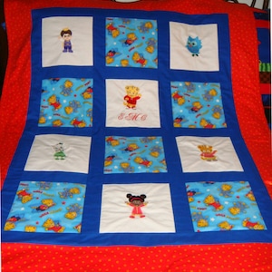 May include: A handmade quilt featuring embroidered cartoon characters and patterns. The quilt has a red border with white polka dots, blue squares, and white squares with embroidered figures. The center square has the embroidered initials "E & MC".