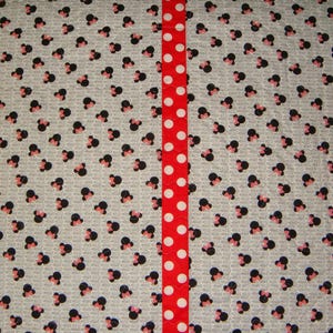 May include: A fabric with a pattern of black and pink Minnie Mouse heads on a light gray background. A vertical red ribbon with white polka dots runs down the center of the fabric.