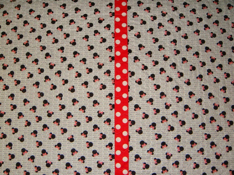 May include: A fabric with a pattern of black and pink Minnie Mouse heads on a light gray background. A vertical red ribbon with white polka dots runs down the center of the fabric.